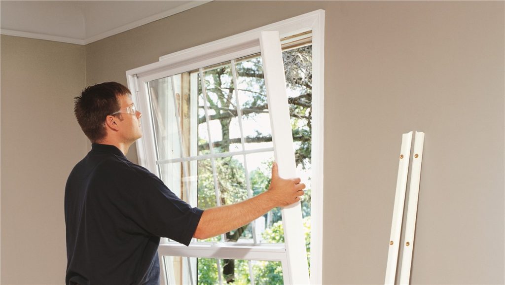 A man wearing glasses and a black shirt is installing or adjusting a white double-hung window inside a room with beige walls.