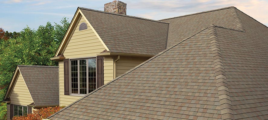 A two-story residential home with yellow siding features a large, multi-angled roof covered in brown and tan architectural shingles, with a stone chimney and dormer windows visible against a partly cloudy sky.